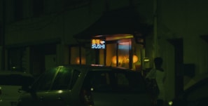 A dimly lit street scene featuring a sushi restaurant with a neon sign. Cars are parked outside, and a person stands near the entrance. Warm yellow and orange lights illuminate the interior from the windows.