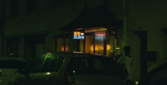 A dimly lit street scene featuring a sushi restaurant with a neon sign. Cars are parked outside, and a person stands near the entrance. Warm yellow and orange lights illuminate the interior from the windows.