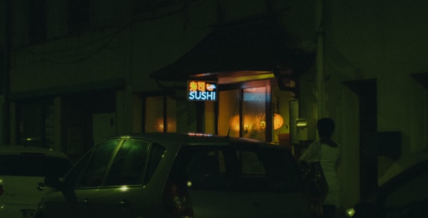 A dimly lit street scene featuring a sushi restaurant with a neon sign. Cars are parked outside, and a person stands near the entrance. Warm yellow and orange lights illuminate the interior from the windows.