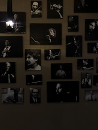 A collection of black and white portraits of musicians is arranged on the wall. Each framed photograph captures a musician either in the midst of playing an instrument or posing with one. The lights add a dramatic shadow, emphasizing the black and white contrasts. The overall atmosphere is artistic and vintage.