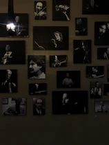 A collection of black and white portraits of musicians is arranged on the wall. Each framed photograph captures a musician either in the midst of playing an instrument or posing with one. The lights add a dramatic shadow, emphasizing the black and white contrasts. The overall atmosphere is artistic and vintage.