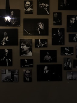 A collection of black and white portraits of musicians is arranged on the wall. Each framed photograph captures a musician either in the midst of playing an instrument or posing with one. The lights add a dramatic shadow, emphasizing the black and white contrasts. The overall atmosphere is artistic and vintage.
