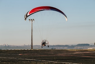 A powered paraglider is in flight close to the ground with a red, black, and white canopy. It is navigating near an open asphalt area with a tall light pole nearby. Distant cityscape and hills are visible under a clear sky.