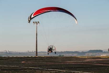 A powered paraglider is in flight close to the ground with a red, black, and white canopy. It is navigating near an open asphalt area with a tall light pole nearby. Distant cityscape and hills are visible under a clear sky.