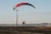 A powered paraglider is in flight close to the ground with a red, black, and white canopy. It is navigating near an open asphalt area with a tall light pole nearby. Distant cityscape and hills are visible under a clear sky.