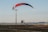 A powered paraglider is in flight close to the ground with a red, black, and white canopy. It is navigating near an open asphalt area with a tall light pole nearby. Distant cityscape and hills are visible under a clear sky.