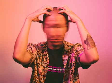 A person with a blurred face, wearing a tropical-patterned shirt and a black shirt underneath with text. They appear to be rubbing or holding their head with both hands. The lighting casts a pinkish hue across the scene, creating a dynamic and slightly distorted visual effect.