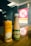 A close-up of two beverage containers on a wooden surface. One is a tall, orange can with Korean text, and the other is a bottle with a beige liquid and green label. The background includes a blurred view of a brightly lit store sign through the glass.