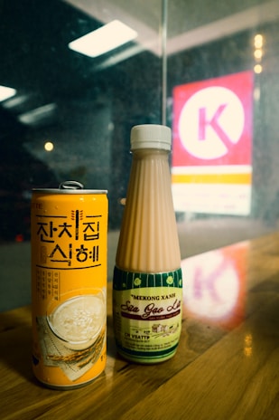 A close-up of two beverage containers on a wooden surface. One is a tall, orange can with Korean text, and the other is a bottle with a beige liquid and green label. The background includes a blurred view of a brightly lit store sign through the glass.