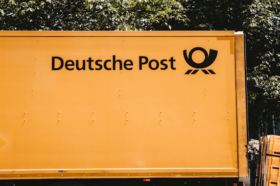 A yellow vehicle with the words 'Deutsche Post' and a postal horn logo on its side. It appears to be a postal or delivery vehicle, parked near some greenery.