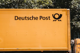 A yellow vehicle with the words 'Deutsche Post' and a postal horn logo on its side. It appears to be a postal or delivery vehicle, parked near some greenery.