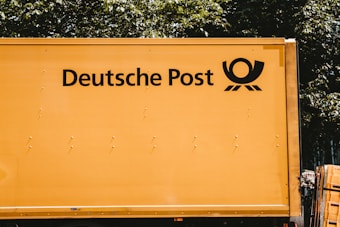A yellow vehicle with the words 'Deutsche Post' and a postal horn logo on its side. It appears to be a postal or delivery vehicle, parked near some greenery.