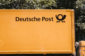 A yellow vehicle with the words 'Deutsche Post' and a postal horn logo on its side. It appears to be a postal or delivery vehicle, parked near some greenery.