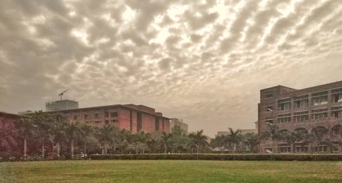 A landscape featuring a campus with several multi-story brick buildings and a large expanse of green lawn in the foreground. Palm trees line the boundaries between the buildings and the lawn. The sky is filled with an impressive display of scattered clouds, giving it a textured appearance. In the background, a construction crane is visible atop one of the buildings, hinting at ongoing development.