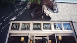 A storefront with windows displaying the words 'ALL DAY BREAKFAST & LUNCH'. Above the windows, there's a vintage-style sign and the exterior is textured and gray. Shadows of tree branches are visible on the wall.