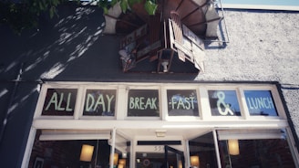 A storefront with windows displaying the words 'ALL DAY BREAKFAST & LUNCH'. Above the windows, there's a vintage-style sign and the exterior is textured and gray. Shadows of tree branches are visible on the wall.
