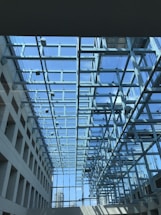 A modern architectural space featuring a glass ceiling supported by a network of metal beams. The clear blue sky is visible through the glass, allowing natural light to flood the interior. The design creates a sense of openness and transparency, with a view of city buildings in the background.