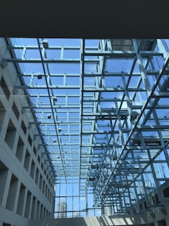 A modern architectural space featuring a glass ceiling supported by a network of metal beams. The clear blue sky is visible through the glass, allowing natural light to flood the interior. The design creates a sense of openness and transparency, with a view of city buildings in the background.