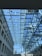 A modern architectural space featuring a glass ceiling supported by a network of metal beams. The clear blue sky is visible through the glass, allowing natural light to flood the interior. The design creates a sense of openness and transparency, with a view of city buildings in the background.