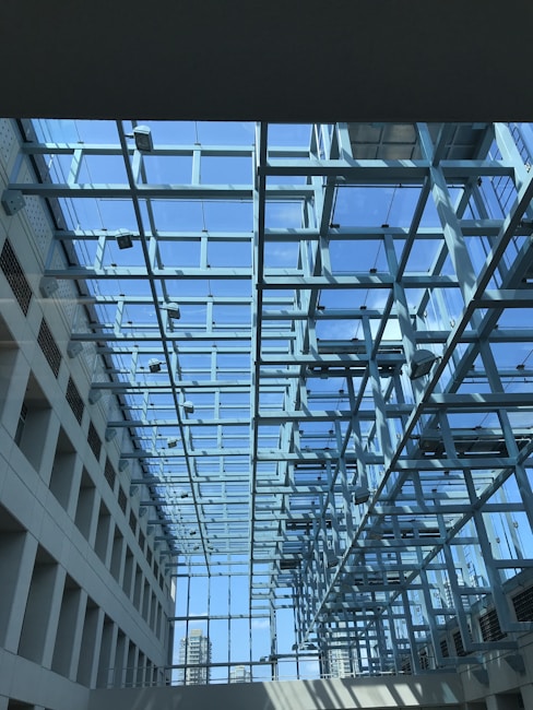 A modern architectural space featuring a glass ceiling supported by a network of metal beams. The clear blue sky is visible through the glass, allowing natural light to flood the interior. The design creates a sense of openness and transparency, with a view of city buildings in the background.