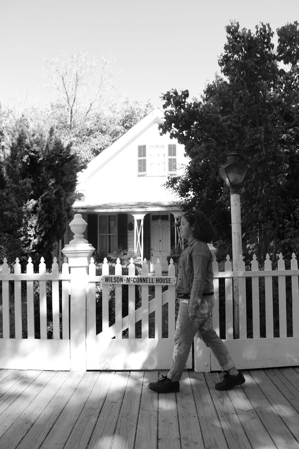 grayscale photo of woman walking beside fence