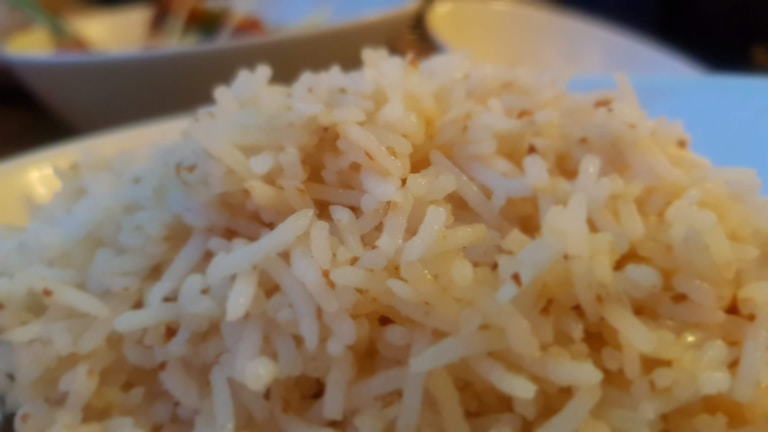 A close-up of steaming white rice, with individual grains clearly visible and slightly fluffy in texture. The background shows blurred hints of dishes with possible vegetables or condiments, providing a warm and cozy dining atmosphere.