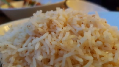 A close-up of steaming white rice, with individual grains clearly visible and slightly fluffy in texture. The background shows blurred hints of dishes with possible vegetables or condiments, providing a warm and cozy dining atmosphere.