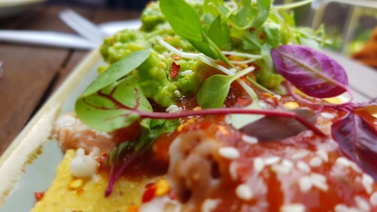 A close-up of a vibrant dish featuring a golden yellow tortilla topped with green guacamole, red sauce, and colorful microgreens. The dish is garnished with sesame seeds and crushed red pepper flakes, creating an appetizing presentation.