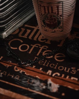 A close-up of a textured paper cup with an intricate logo sits atop a vintage-style coffee sign featuring decorative typography. There is a metal chain draped across the surface, adding a subtle industrial touch. The lighting is dim, casting warm shadows and emphasizing the retro ambiance.