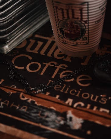 A close-up of a textured paper cup with an intricate logo sits atop a vintage-style coffee sign featuring decorative typography. There is a metal chain draped across the surface, adding a subtle industrial touch. The lighting is dim, casting warm shadows and emphasizing the retro ambiance.