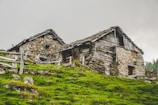Two rustic stone and wooden houses are nestled on a grassy hillside. The buildings exhibit an aged, quaint appearance with weathered wooden roofs and stone walls. A wooden fence surrounds the area, adding to the rural setting. The sky is overcast, lending a muted atmosphere to the scene.