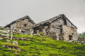 Two rustic stone and wooden houses are nestled on a grassy hillside. The buildings exhibit an aged, quaint appearance with weathered wooden roofs and stone walls. A wooden fence surrounds the area, adding to the rural setting. The sky is overcast, lending a muted atmosphere to the scene.