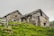 Two rustic stone and wooden houses are nestled on a grassy hillside. The buildings exhibit an aged, quaint appearance with weathered wooden roofs and stone walls. A wooden fence surrounds the area, adding to the rural setting. The sky is overcast, lending a muted atmosphere to the scene.