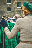 A person with a retro hairstyle is wearing a green beret and a checkered coat, standing in front of a rack of vintage green dresses with a lively pattern. The setting appears to be a market or vintage fair, with additional blurred figures in the background and a brick building behind.