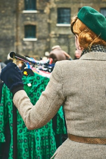 A person with a retro hairstyle is wearing a green beret and a checkered coat, standing in front of a rack of vintage green dresses with a lively pattern. The setting appears to be a market or vintage fair, with additional blurred figures in the background and a brick building behind.