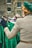 A person with a retro hairstyle is wearing a green beret and a checkered coat, standing in front of a rack of vintage green dresses with a lively pattern. The setting appears to be a market or vintage fair, with additional blurred figures in the background and a brick building behind.