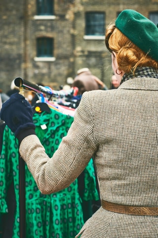 A person with a retro hairstyle is wearing a green beret and a checkered coat, standing in front of a rack of vintage green dresses with a lively pattern. The setting appears to be a market or vintage fair, with additional blurred figures in the background and a brick building behind.