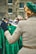 A person with a retro hairstyle is wearing a green beret and a checkered coat, standing in front of a rack of vintage green dresses with a lively pattern. The setting appears to be a market or vintage fair, with additional blurred figures in the background and a brick building behind.