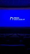 A dimly lit movie theater featuring rows of empty seats silhouetted against a large blue screen displaying the logo and website of the Institute of Cinema and Audiovisual Arts (INCAA) in Argentina.