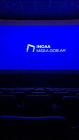 A dimly lit movie theater featuring rows of empty seats silhouetted against a large blue screen displaying the logo and website of the Institute of Cinema and Audiovisual Arts (INCAA) in Argentina.