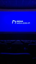 A dimly lit movie theater featuring rows of empty seats silhouetted against a large blue screen displaying the logo and website of the Institute of Cinema and Audiovisual Arts (INCAA) in Argentina.