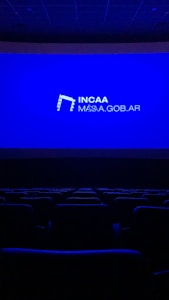 A dimly lit movie theater featuring rows of empty seats silhouetted against a large blue screen displaying the logo and website of the Institute of Cinema and Audiovisual Arts (INCAA) in Argentina.