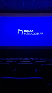 A dimly lit movie theater featuring rows of empty seats silhouetted against a large blue screen displaying the logo and website of the Institute of Cinema and Audiovisual Arts (INCAA) in Argentina.