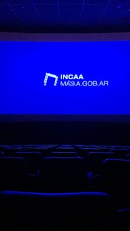 A dimly lit movie theater featuring rows of empty seats silhouetted against a large blue screen displaying the logo and website of the Institute of Cinema and Audiovisual Arts (INCAA) in Argentina.