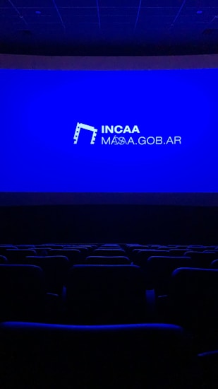 A dimly lit movie theater featuring rows of empty seats silhouetted against a large blue screen displaying the logo and website of the Institute of Cinema and Audiovisual Arts (INCAA) in Argentina.