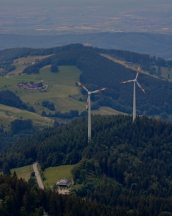 Two large wind turbines are situated in a lush, forested area with hills and scattered clearings. A small road winds through the greenery, leading to a building nestled among the trees. The landscape features expansive views of rolling hills, fields, and forested areas.