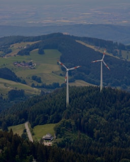 Two large wind turbines are situated in a lush, forested area with hills and scattered clearings. A small road winds through the greenery, leading to a building nestled among the trees. The landscape features expansive views of rolling hills, fields, and forested areas.
