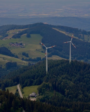 Two large wind turbines are situated in a lush, forested area with hills and scattered clearings. A small road winds through the greenery, leading to a building nestled among the trees. The landscape features expansive views of rolling hills, fields, and forested areas.