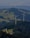 Two large wind turbines are situated in a lush, forested area with hills and scattered clearings. A small road winds through the greenery, leading to a building nestled among the trees. The landscape features expansive views of rolling hills, fields, and forested areas.