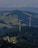 Two large wind turbines are situated in a lush, forested area with hills and scattered clearings. A small road winds through the greenery, leading to a building nestled among the trees. The landscape features expansive views of rolling hills, fields, and forested areas.